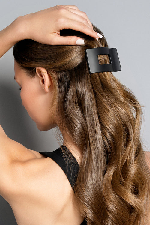 A brunette model with long, wavy hair wearing a black rectangle flat lay claw clip against a gray background