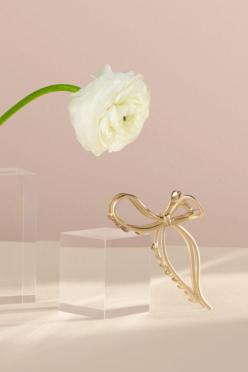 The Hair Edit Gold Gilded Bow Claw Clip in a lifestyle setting with a white flower