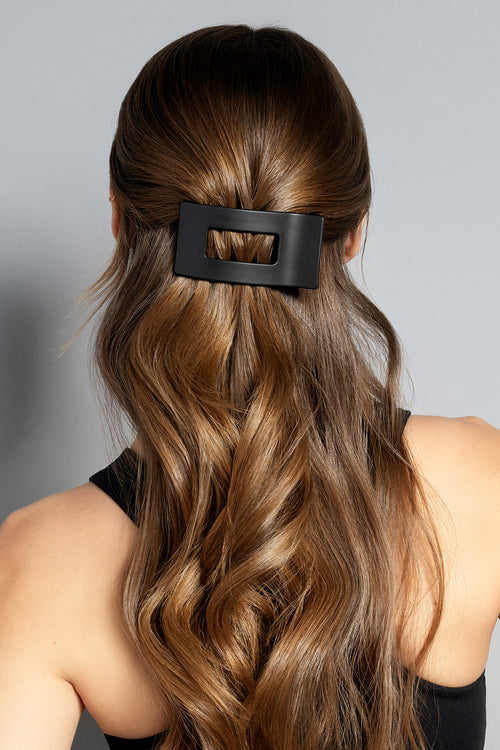 A brunette model with long wavy hair wearing a black rectangle flat lay claw clip on a gray background