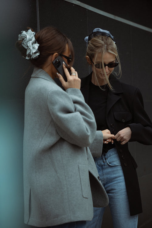 Two women standing against a dark wall, one using a phone and the other looking at something wearing The Hair Edit's oversized scrunchies